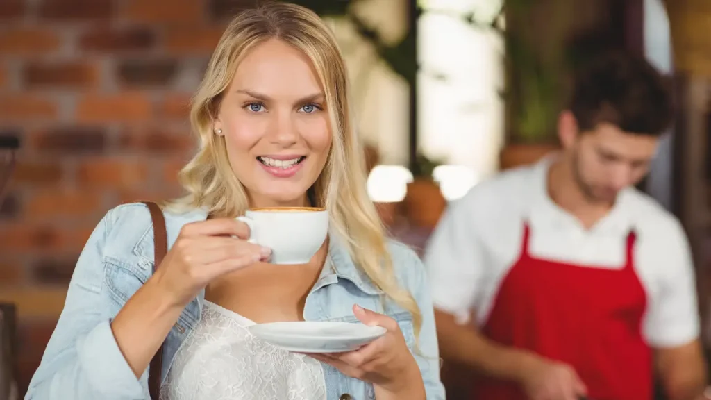 Cliente de cafeteria tomando café em xícara com um sorriso e ao fundo movimento com atendente